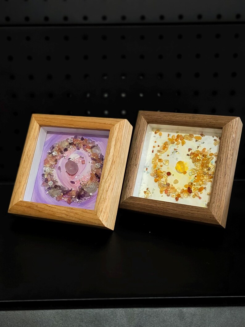 Pu&ograve; includere: Due opere d'arte incorniciate con cornici in legno. Una presenta un design viola e rosa con cristalli, l'altra ha una disposizione di cristalli arancioni e gialli. Entrambe sono quadrate e poste su uno sfondo scuro.
