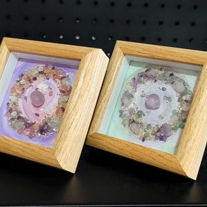 Pu&ograve; includere: Due opere d'arte incorniciate con cornici in legno chiaro. Ognuna presenta una pietra rosa centrale circondata da una disposizione circolare di cristalli e pietre colorate. Gli sfondi sono dipinti con colori pastello tenui.