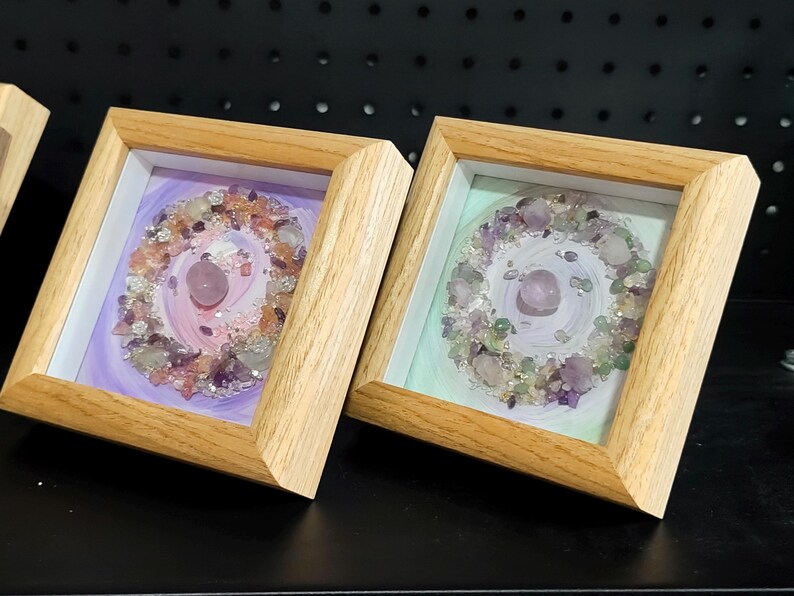Pu&ograve; includere: Due opere d'arte incorniciate con cornici in legno chiaro. Ognuna presenta una pietra rosa centrale circondata da una disposizione circolare di cristalli e pietre colorate. Gli sfondi sono dipinti con colori pastello tenui.
