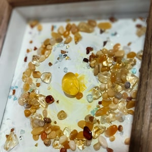 Pu&ograve; includere: Un'opera d'arte incorniciata con una collezione di pietre preziose lucide nei toni del giallo, arancione e trasparente. Le pietre sono disposte su una superficie bianca con un centro giallo, all'interno di una cornice di legno.