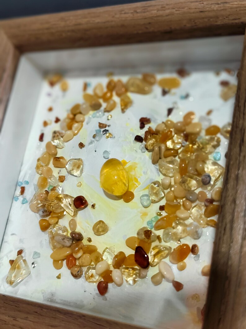 Pu&ograve; includere: Un'opera d'arte incorniciata con una collezione di pietre preziose lucide nei toni del giallo, arancione e trasparente. Le pietre sono disposte su una superficie bianca con un centro giallo, all'interno di una cornice di legno.