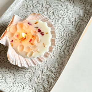 Peut inclure: Une bougie allumée dans une coquille Saint-Jacques naturelle, contenant de la cire blanche incrustée de pétales de fleurs roses et orange séchés. La coquille repose sur un plateau rectangulaire blanc au motif de feuilles et de vignes en relief.
