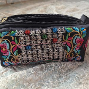 Peut inclure: Petite pochette rectangulaire noire avec fermeture éclair et ruban noir. La pochette est ornée de broderies florales colorées roses, bleues, vertes et jaunes. La broderie comprend un motif de formes blanches en forme de fleurs.