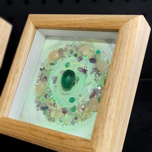 Pu&ograve; includere: Un'opera d'arte incorniciata con un design circolare, caratterizzata da una pietra verde centrale. Il design &egrave; circondato da piccole gemme colorate e dettagli argentati, su uno sfondo verde chiaro. La cornice &egrave; in legno chiaro.