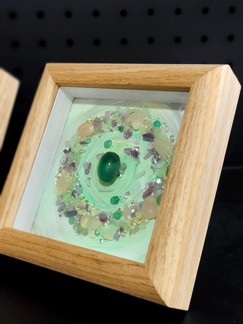 Pu&ograve; includere: Un'opera d'arte incorniciata con un design circolare, caratterizzata da una pietra verde centrale. Il design &egrave; circondato da piccole gemme colorate e dettagli argentati, su uno sfondo verde chiaro. La cornice &egrave; in legno chiaro.