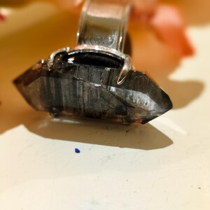 May include: A silver ring with a dark grey, faceted crystal. The crystal is pointed and set in a silver band. The background includes soft pink and orange flower petals.