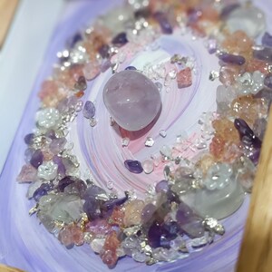 Pu&ograve; includere: Un'opera d'arte incorniciata con una pietra centrale liscia color lavanda circondata da una spirale di pietre preziose. Le pietre preziose includono ametista, quarzo trasparente e cristalli rosa e arancioni, su uno sfondo dipinto di lavanda e bianco.