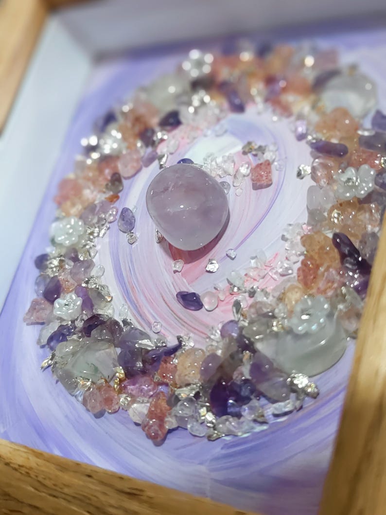 Pu&ograve; includere: Un'opera d'arte incorniciata con una pietra centrale liscia color lavanda circondata da una spirale di pietre preziose. Le pietre preziose includono ametista, quarzo trasparente e cristalli rosa e arancioni, su uno sfondo dipinto di lavanda e bianco.