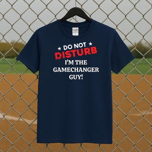 Könnte beinhalten: Marineblaues T-Shirt mit dem roten Text "DO NOT DISTURB" und dem weißen Text "I'M THE GAMECHANGER GUY!". Das T-Shirt hängt an einem schwarzen Kleiderbügel vor einem Maschendrahtzaun und einem Baseballfeld.