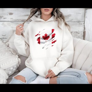 Op de afbeelding: Crèmekleurige hoodie met een versleten Canadees vlagontwerp. De rode en witte kleuren van de vlag zijn zichtbaar door het gescheurde stofeffect. De hoodie heeft een voorzak en een capuchon met trekkoord.