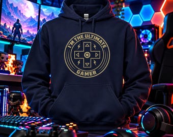May include: A navy blue hooded sweatshirt featuring a gold circular graphic on the chest. The design includes a central directional pad icon surrounded by the text I'M THE ULTIMATE GAMER, with small lightning bolt symbols on the sides.
