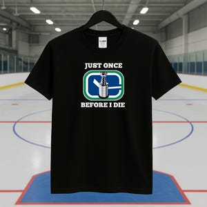 May include: Black t-shirt featuring the text "JUST ONCE BEFORE I DIE" above and below a hockey trophy graphic within a blue and green rectangle. The shirt is displayed on a black hanger.