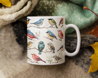 May include: White ceramic mug with a colorful pattern of birds on branches. The mug has a white handle and is sitting on a brown and green blanket with yellow and orange leaves.
