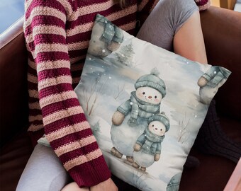 Op de afbeelding: Een wit kussen met een aquarel-illustratie van twee sneeuwpoppen met blauwe sjaals en hoeden. De sneeuwpoppen staan in een besneeuwd landschap met bomen en sneeuwvlokken.