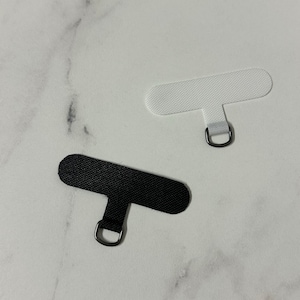 May include: Two phone lanyard patches, one black and one white, each with a small metal D-ring. The patches are rectangular with a smaller tab and ring for attachment. The background is a white marble surface.