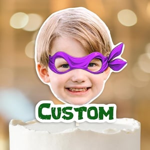 May include: A white cake with blue frosting and a custom cake topper featuring a child wearing a purple ninja turtle mask. The topper says "CUSTOM" in green letters.