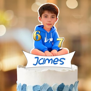 Puede incluir: Una tarta de cumpleaños con glaseado blanco y detalles decorativos azules. Un adorno para tarta presenta una imagen de dibujos animados de un niño con una sudadera con capucha azul, con los números 6 y 7. El nombre "James" está escrito en una pancarta.