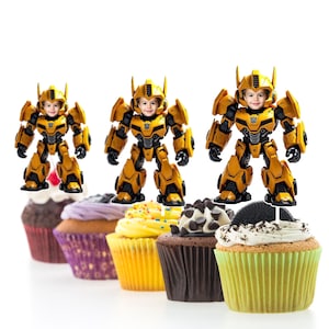 May include: Yellow and black robot cupcake toppers with a child's face on the robot's head. The toppers are on top of cupcakes with different colored frosting.