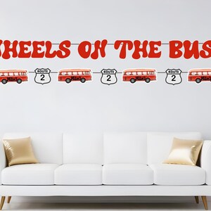 May include: A red and white banner with the words "WHEELS ON THE BUS" in large, red, stylized letters. The banner is decorated with red and white stop signs, red school buses, and route signs.