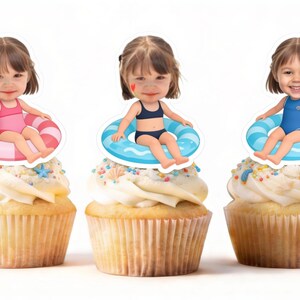 May include: Three cupcakes topped with custom paper cutouts featuring a child's face on a cartoon body wearing a swimsuit and sitting in a colorful inflatable pool ring. The cupcakes have white frosting decorated with small colorful sprinkles.