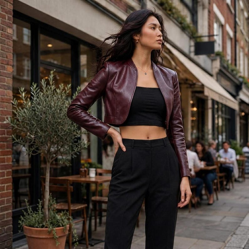 A model standing on a city street wearing a burgundy leather cropped jacket over a black crop top and black trousers.