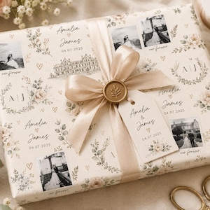 May include: A rectangular gift wrapped in cream-colored paper featuring floral illustrations, black and white wedding photographs, the names Amelia and James, the date 04.07.2025, and a beige satin ribbon tied with a gold wax seal and a matching gift tag.