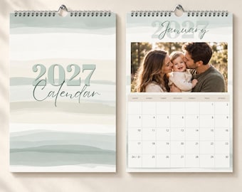May include: A 2027 wall calendar with a spiral binding, featuring a cover with abstract sage green and beige watercolor stripes and the text 2027 Calendar. The open page shows a photograph of a family and a January 2027 grid calendar.