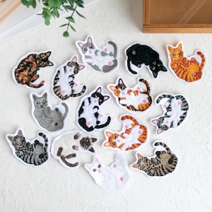 以下が含まれることがあります： 様々な色と模様の刺繍された猫のパッチのコレクション。パッチは、縞模様、斑点、さまざまな毛の色などの詳細を備えた、遊び心のあるポーズの猫を特徴としています。パッチは白い表面にあります。