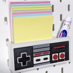 May include: A white 3D printed Nintendo controller wall organiser with a black and grey design. The controller has a slot for holding notes or papers. The organiser is mounted on a white pegboard wall. There is a small bottle of glue in the slot.