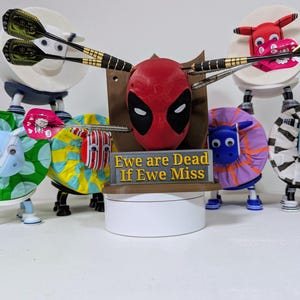 May include: A whimsical dartboard display featuring a Deadpool mask, surrounded by sheep-shaped targets. The targets are decorated with various patterns and colors, and the sign reads "Ewe are Dead If Ewe Miss". Darts are positioned as if thrown.