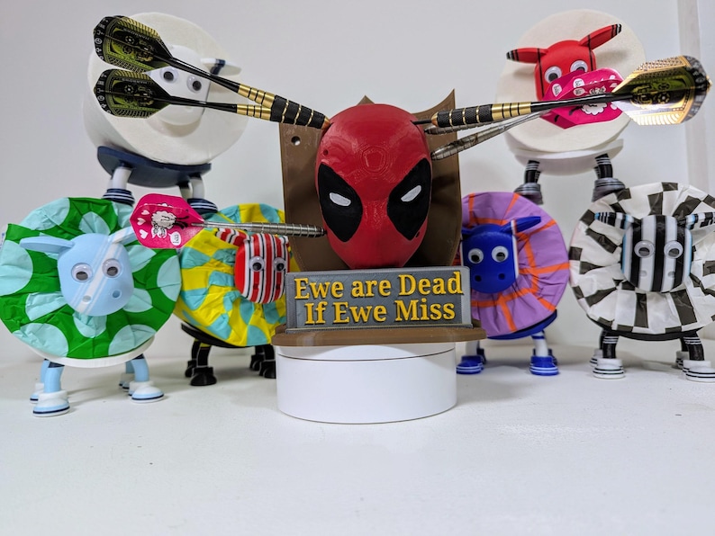 May include: A whimsical dartboard display featuring a Deadpool mask, surrounded by sheep-shaped targets. The targets are decorated with various patterns and colors, and the sign reads "Ewe are Dead If Ewe Miss". Darts are positioned as if thrown.