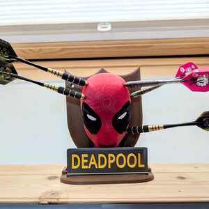 May include: A Deadpool-themed dartboard featuring a red mask with black eyes and a brown plaque. Darts are embedded in the mask. The base reads "DEADPOOL" in yellow letters.