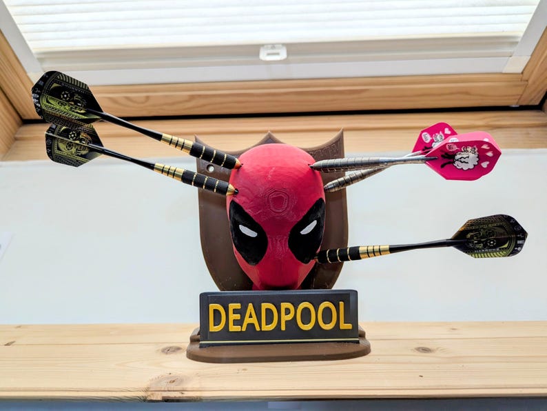 May include: A Deadpool-themed dartboard featuring a red mask with black eyes and a brown plaque. Darts are embedded in the mask. The base reads "DEADPOOL" in yellow letters.