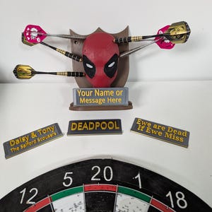 May include: A Deadpool-themed dartboard set. The centerpiece is a red Deadpool mask with darts embedded. Includes a dartboard, nameplates with text, and a sign that reads "Your Name or Message Here."