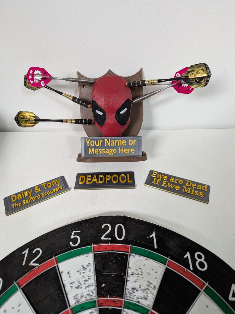 May include: A Deadpool-themed dartboard set. The centerpiece is a red Deadpool mask with darts embedded. Includes a dartboard, nameplates with text, and a sign that reads "Your Name or Message Here."