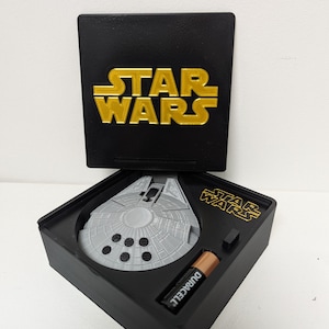 May include: A black box featuring the gold text "STAR WARS" on the lid. Inside, a grey Millennium Falcon model is displayed in a black base, alongside a Duracell battery. The Star Wars logo is also on the base.