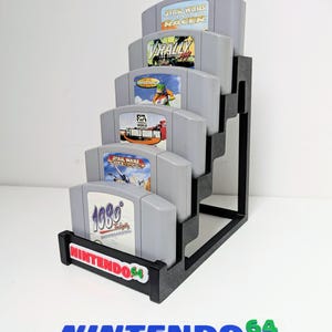 May include: A black stepped display stand holding five grey Nintendo 64 game cartridges. The games include Star Wars Episode I Racer, V-Rally Edition, Wave Race 64, F-1 World Grand Prix, and 1080° Snowboarding. The stand has the text "NINTENDO 64".
