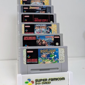 Può includere: Una pila di cartucce di giochi per Super Nintendo, tra cui "Super Mario All-Stars" e "Yoshi's Safari". Le cartucce grigie presentano illustrazioni colorate e testo. Un supporto bianco con "SUPER FAMICOM" è alla base.