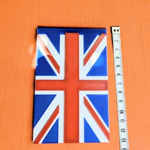 May include: A rectangular Union Jack flag plaque featuring raised red, white, and blue sections. A white measuring tape is positioned vertically next to the plaque, showing a length of approximately 33 cm for the object's vertical side.