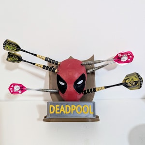 May include: A Deadpool-themed dartboard featuring a red mask with black eye cutouts, surrounded by darts. The base reads "DEADPOOL" in yellow letters. The darts have black, gold, and pink accents.