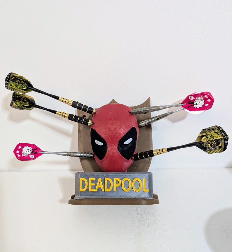 May include: A Deadpool-themed dartboard featuring a red mask with black eye cutouts, surrounded by darts. The base reads "DEADPOOL" in yellow letters. The darts have black, gold, and pink accents.