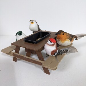 Könnte beinhalten: Ein kleiner brauner Picknicktisch mit einem schwarzen Behälter in der Mitte, umgeben von vier dekorativen Vogelfiguren. Die Vögel sind in verschiedenen Farben gehalten, darunter Weiß, Grün, Rot und Braun, mit detaillierten Federn.