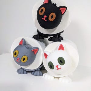 Op de afbeelding: Drie 3D-geprinte kattenvormige toiletpapierhouders. De bovenste houder is zwart met oranje ogen, de middelste houder is grijs met gele ogen en de onderste houder is wit met groene ogen.
