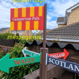 May include: A brown plastic signpost featuring a rectangular flag with red and yellow vertical stripes and the text Campervan Pals. Below the flag, four directional arrows point outward, labeled Wales, Northern Ireland, England, and Scotland.