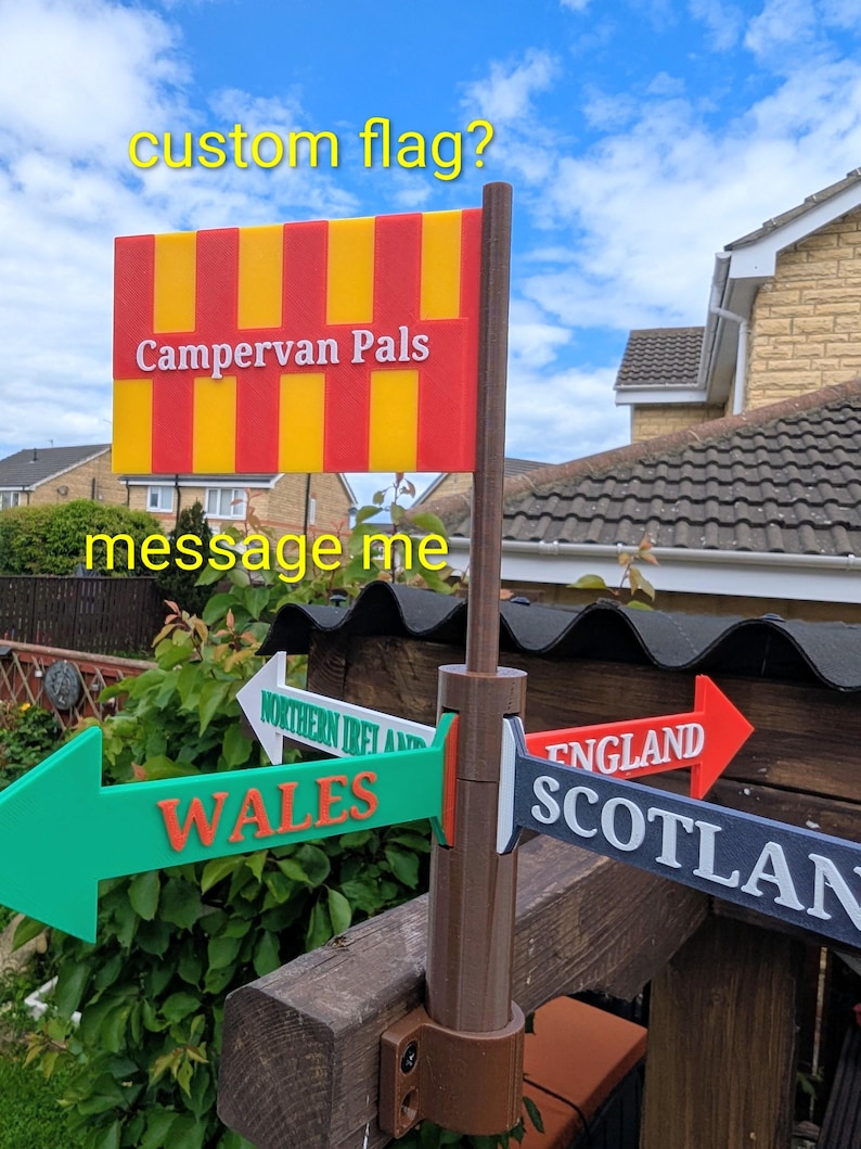 May include: A brown plastic signpost featuring a rectangular flag with red and yellow vertical stripes and the text Campervan Pals. Below the flag, four directional arrows point outward, labeled Wales, Northern Ireland, England, and Scotland.