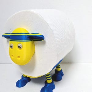 Può includere: Un portarotolo di carta igienica stravagante a forma di pecora. La pecora ha una testa gialla con dettagli a strisce blu e gialle, piedi blu e zampe a righe gialle e blu. Il rotolo di carta igienica forma il corpo della pecora.