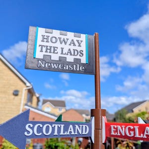 May include: A brown plastic signpost featuring a top rectangular sign with white text reading HOWAY THE LADS and Newcastle, and two lower directional arrow signs pointing left and right, labelled SCOTLAND and ENGLA in white raised lettering.
