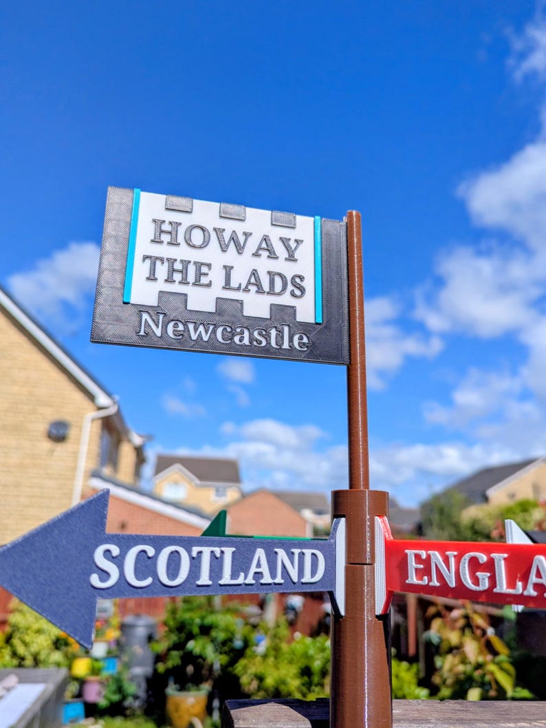 May include: A brown plastic signpost featuring a top rectangular sign with white text reading HOWAY THE LADS and Newcastle, and two lower directional arrow signs pointing left and right, labelled SCOTLAND and ENGLA in white raised lettering.