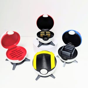 Può includere: Quattro contenitori sferici aperti, rossi, gialli e blu con dettagli neri. I contenitori hanno basi bianche e gambe grigie. Uno dei contenitori è aperto, rivelando un oggetto dorato.