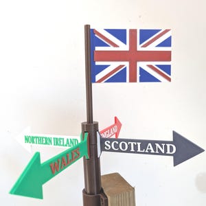 May include: A brown plastic signpost featuring a Union Jack flag at the top and four directional arrows pointing outward. The arrows are labelled with the text Northern Ireland, Wales, England, and Scotland in various colours and fonts.